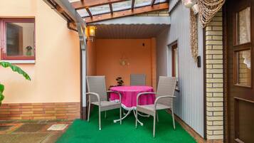 Een overdekt terras met een tafel, stoelen, een deur en een muur met een decoratieve wandhanger.