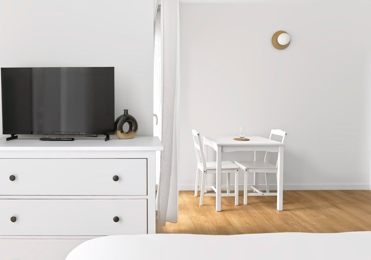 Una habitación minimalista con un tocador blanco, un televisor, una mesita con sillas y un adorno redondo en la pared.