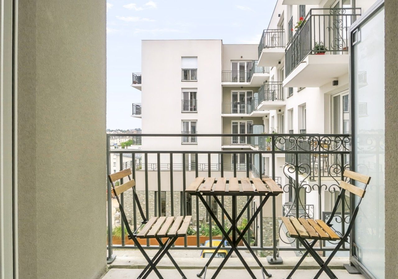 Un balcon doté de deux tables et de deux chaises en bois, d’une rambarde en métal et offrant une vue sur les bâtiments adjacents.