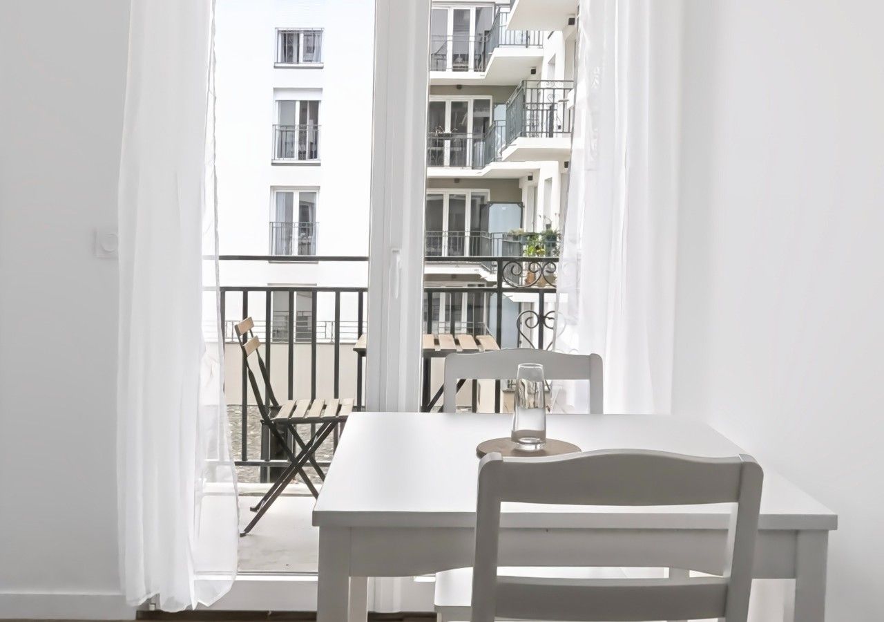 Un balcón con una mesa y sillas blancas, un vaso de agua y vistas a un edificio de varios pisos.