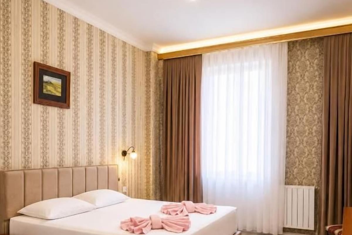 A hotel room with a large bed, a bedside table, a nightstand, a lamp, and a window with curtains.