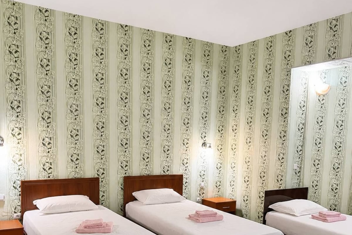 A hotel room with three single beds, a green carpet with a leaf pattern, and ornate wallpaper.
