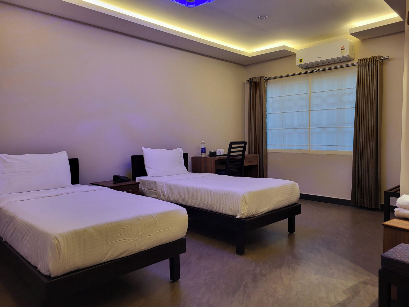 A hotel room with two beds, a desk, a chair, an air conditioning unit, and a window with curtains.