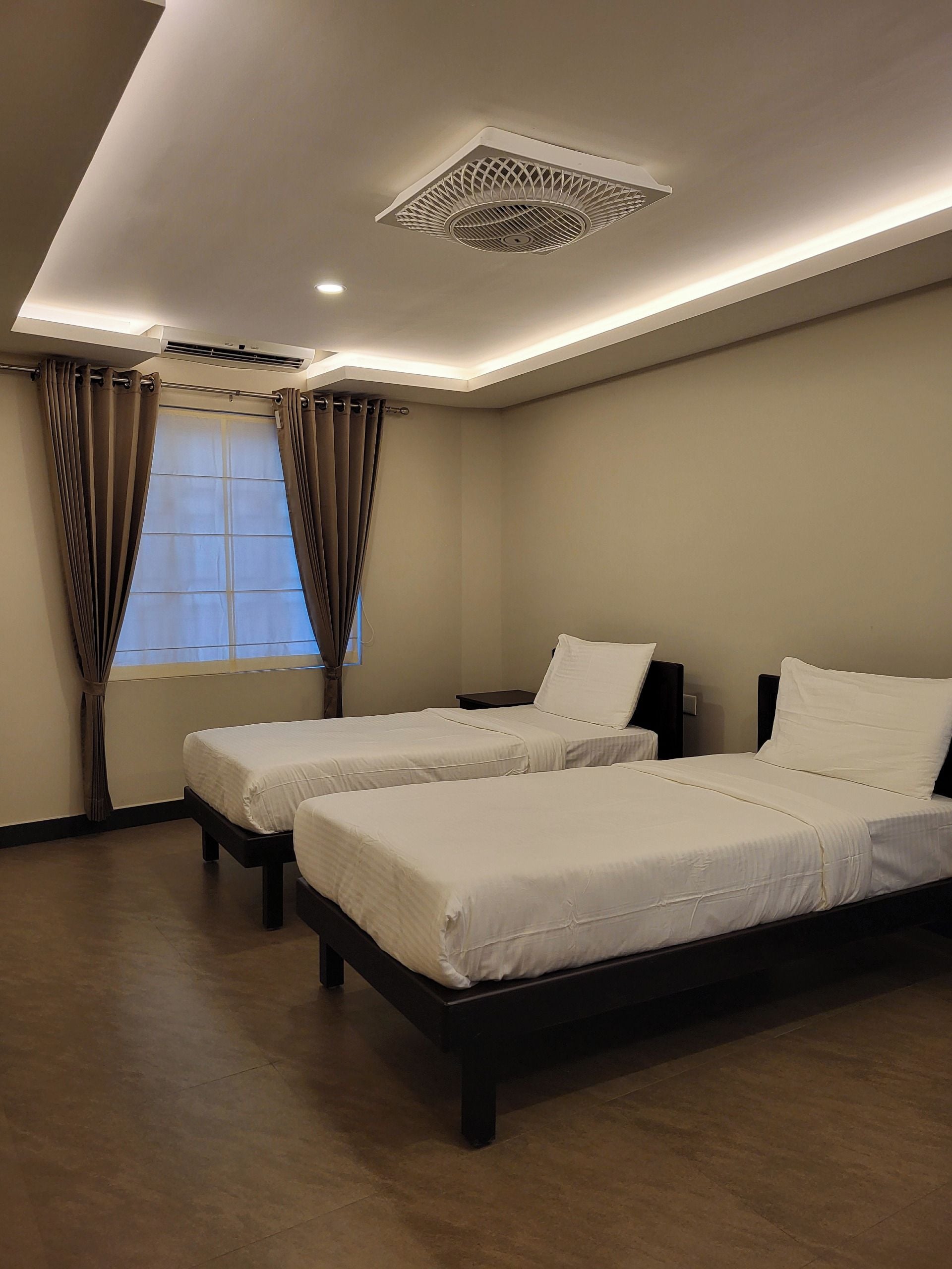 A hotel room with two beds, a window with curtains, a ceiling vent, and a wall-mounted air conditioning unit.