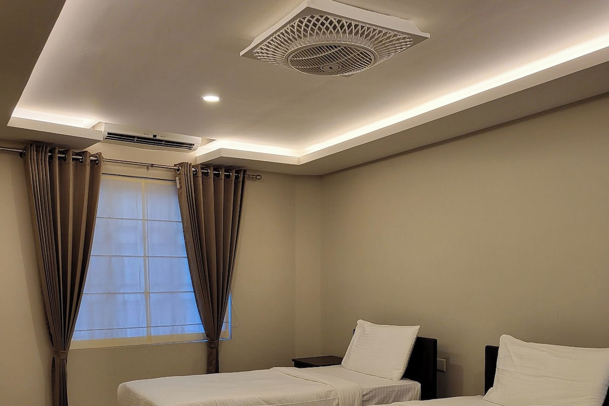 Habitación de hotel con dos camas, una ventana con cortinas, un respiradero en el techo y un aire acondicionado instalado en la pared.
