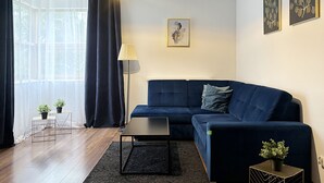 Uma sala de estar moderna com um sofá azul escuro, uma mesa de centro preta, um abajur e uma janela grande com cortinas leves.