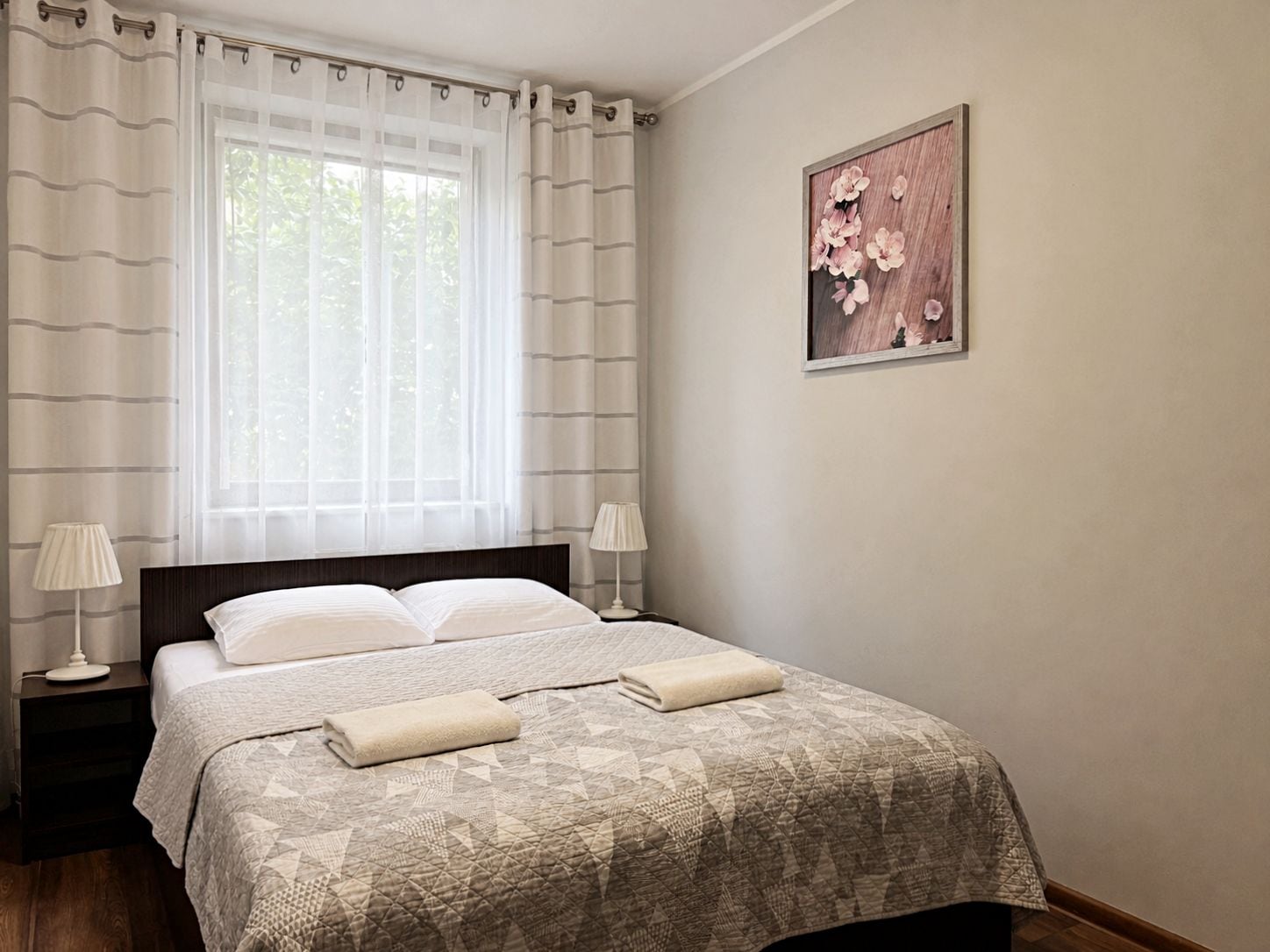 Ein Hotelzimmer mit einem Bett, zwei Nachttischlampen, einem fensterbelaubten Fenster und einem Wandbild.