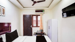A hotel room with a bed, a ceiling fan, an air conditioner, a refrigerator, a small table with a kettle, and a window with curtains. - Townhouse Emerald Mall Lucknow (Lucknow)