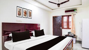 A hotel room with a bed, a nightstand, a desk with a kettle, a refrigerator, a window with bars, and a ceiling fan. - Townhouse Emerald Mall Lucknow (Lucknow)