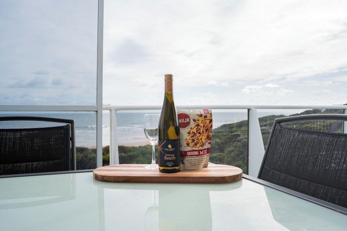 A table with a bottle of wine, a glass, and a box of snacks on a balcony overlooking a scenic view.