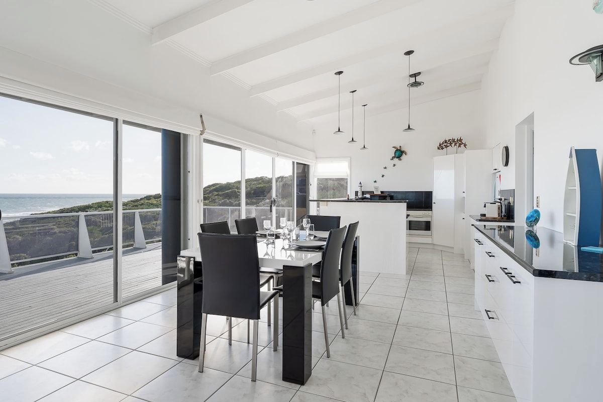A modern kitchen with a large island, a dining area with a table and chairs, and a view of the sea through large windows.