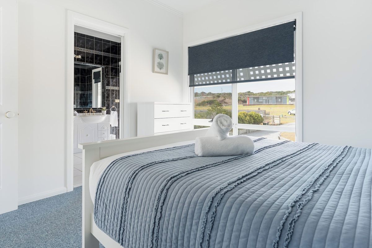 A bedroom with a bed, a white wardrobe, a bathroom with a sink, and a window with a view of buildings.