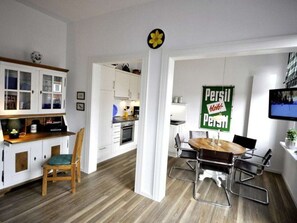 Eine moderne Küche mit weißen Schränken, einem Holztisch mit Metallstühlen und einem an der Wand montierten Fernseher.