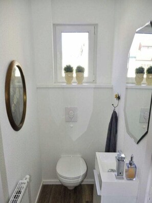 Un bagno piccolo e bianco con il water, un lavandino, uno specchio e una finestra con piante in vaso.