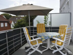 A balcony with a table and chairs, an umbrella, and a view of buildings and trees. - Holiday Apartment Napoleonschanze (Norderney)
