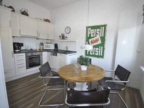 A compact kitchen with white cabinets, a wooden dining table with four chairs, and a Persil logo on the wall. - Holiday Apartment Napoleonschanze (Norderney)