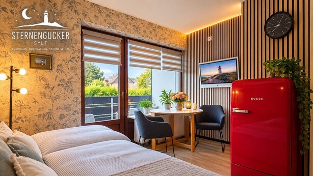 A hotel room with a bed, a red Bosch refrigerator, a dining table with chairs, a TV on the wall, and a window with blinds. - Apartment 'Sternengucker Sylt' with Balcony and Wi-Fi (Wenningstedt-Braderup (Sylt))