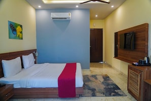 Habitación de hotel con cama, televisor montado en la pared, cabecero de madera, mesita de noche, manta roja, un cuadro y una puerta que da a otra habitación.