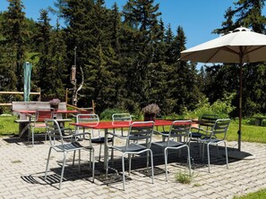 En udendørs terrasse med et rødt bord og metalstole, omgivet af grøn vegetation og træer.