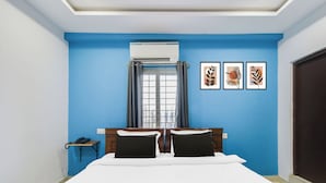 Une chambre d’hôtel avec un lit, une fenêtre avec des rideaux, un climatiseur fixé au mur et trois œuvres d’art encadrées.