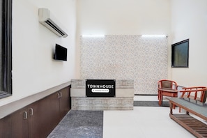Zona de recepción con un cartel que indica que es un lugar de servicio de empresa, un televisor de pantalla plana, un muro con diseño y un área de estar con un banco y sillas.