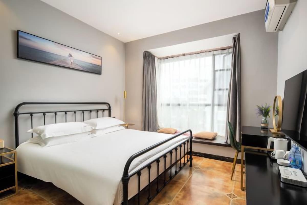 Un dormitorio con cama, escritorio, silla, ventana con cortinas y un cuadro en la pared.