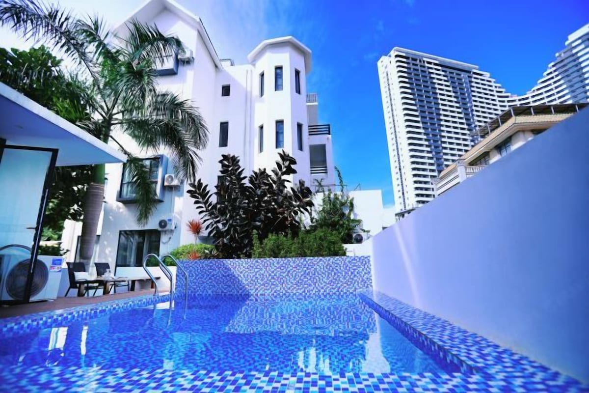 Una piscina con piso de baldosas, rodeada de edificios modernos y follaje.