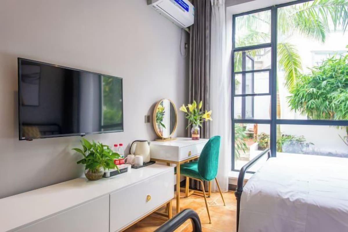 Habitación de hotel con cama, escritorio y silla, televisor y ventana con vista a follaje.