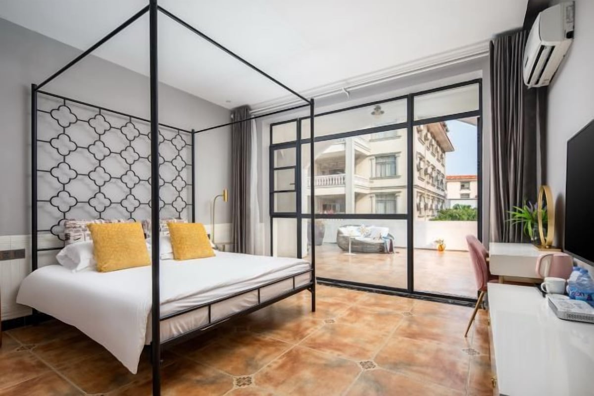 Habitación con cama con dosel, un escritorio con silla y un amplio ventanal con vistas a edificios.