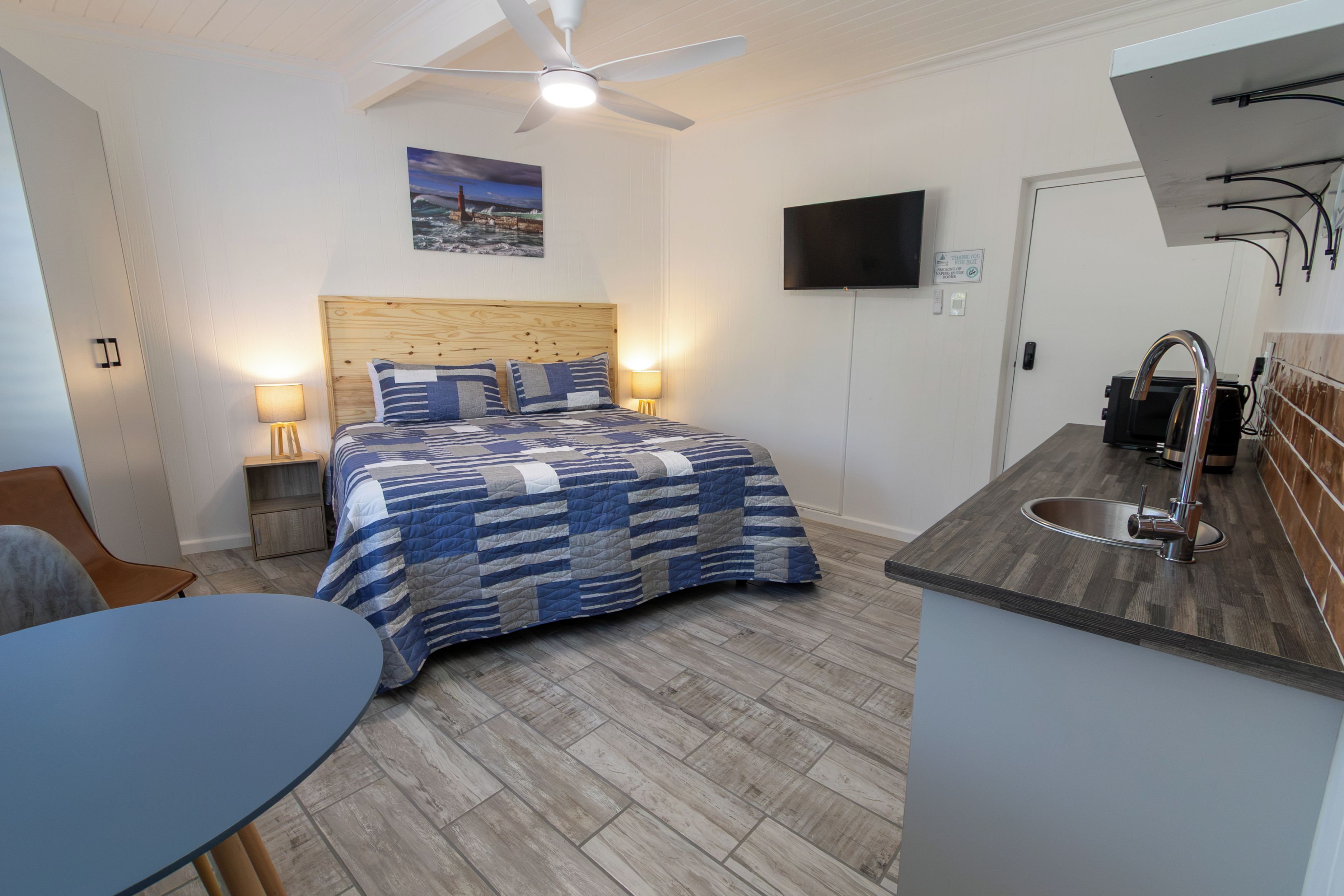 A bedroom with a bed, bedside lamps, a ceiling fan, a TV, and a kitchenette with a sink.