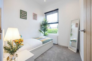 Ein Schlafzimmer mit Bett, Nachttischlampe, Heizkörper, einem Fenster mit Jalousien und motivierender Wandkunst.