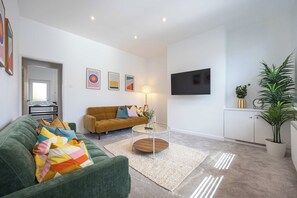 Een moderne woonkamer met een groene bank, een mosterdgele bank en een salontafel. Er is een flatscreen televisie aan de muur en een kamerplant aan de rechterkant.