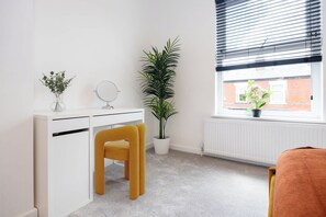Een kleine, netjes ingerichte kamer met een wit bureau, een spiegel, een gele stoel, een kamerplant en een raam met jaloezieën.