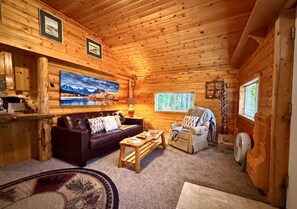 A log cabin interior with a wooden ceiling, a large painting, a sofa, a chair, a table, and a fan. - Peaceful Cabin with Fire Pit & Endless Trail Adventures in Island Park, Idaho (Island Park)