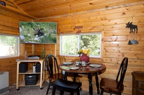 A wooden cabin interior with a dining table set for four, a television mounted on the wall, and a window with a view of greenery. - Peaceful Cabin with Fire Pit & Endless Trail Adventures in Island Park, Idaho (Island Park)