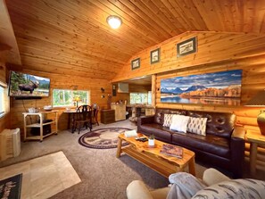 L’intérieur chaleureux d’un chalet avec un plafond en bois, un canapé en cuir, un coin repas et une grande fenêtre donnant sur un paysage magnifique.