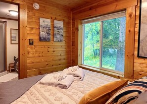 Une chambre dans un chalet en bois, comprenant un lit, deux serviettes, une fenêtre donnant sur la forêt et une petite table avec une chaise.
