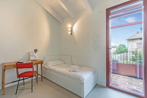 A small, single-bed room with a desk and chair, a window with a view of buildings, and a ceiling with exposed beams. - Molinella Easy Living Apartment (Modena)