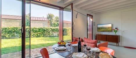 Ein Essbereich mit einem Tisch für zwei Personen, einem Fernseher an der Wand und Blick auf einen Garten durch große Glasüren.