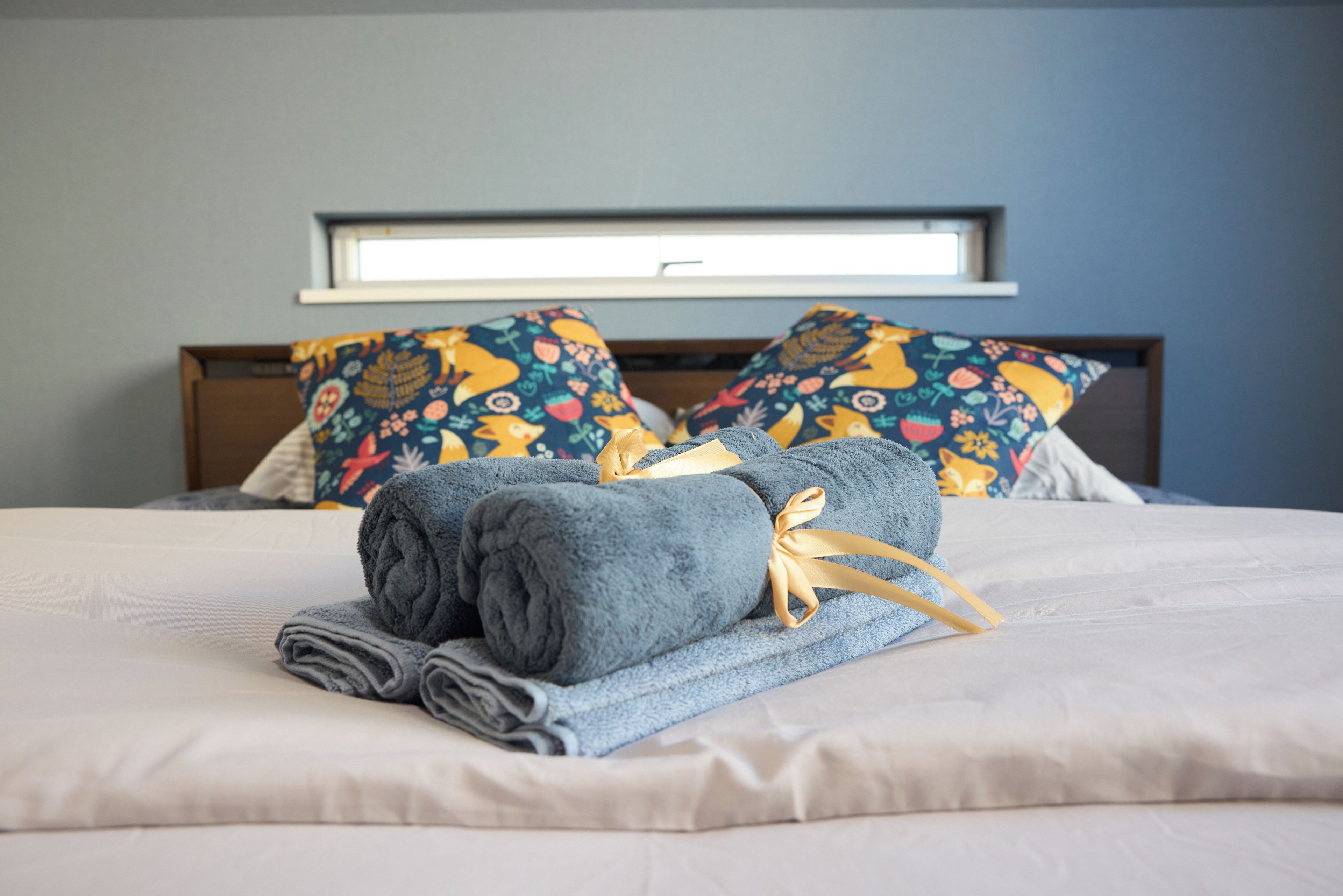 Cama bem arrumada com toalhas enroladas e uma cesta de presentes, em frente a uma parede azul com almofadas estampadas.