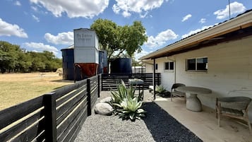 Un bâtiment sur lequel est apposée une enseigne indiquant « Big Poshman Store-Vey », un espace extérieur clôturé avec un banc, une table et des chaises, ainsi qu’un chemin de gravier.