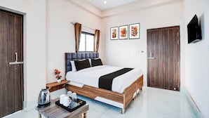 Quarto de hotel moderno com uma cama grande, mesa de cabeceira com chaleira e xícaras, TV fixada na parede e uma janela com cortinas.