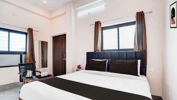 Quarto de hotel com cama, escrivaninha com chaleira, cadeira, janela com cortinas e espelho fixado na parede.