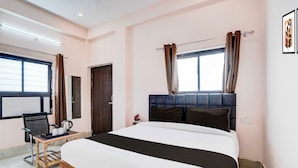 Quarto de hotel com uma cama grande, mesa de cabeceira de madeira, uma cadeira e janela com cortinas.