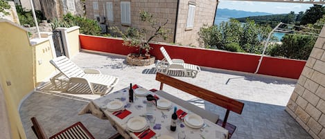 An outdoor patio with a table set for dining, surrounded by lounge chairs and a view of the sea and mountains.