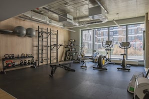 Ein Fitnessraum mit unterschiedlicher Trainingsausstattung, einer Holzvertäfelung und einem großen Fenster mit Blick auf Gebäude.