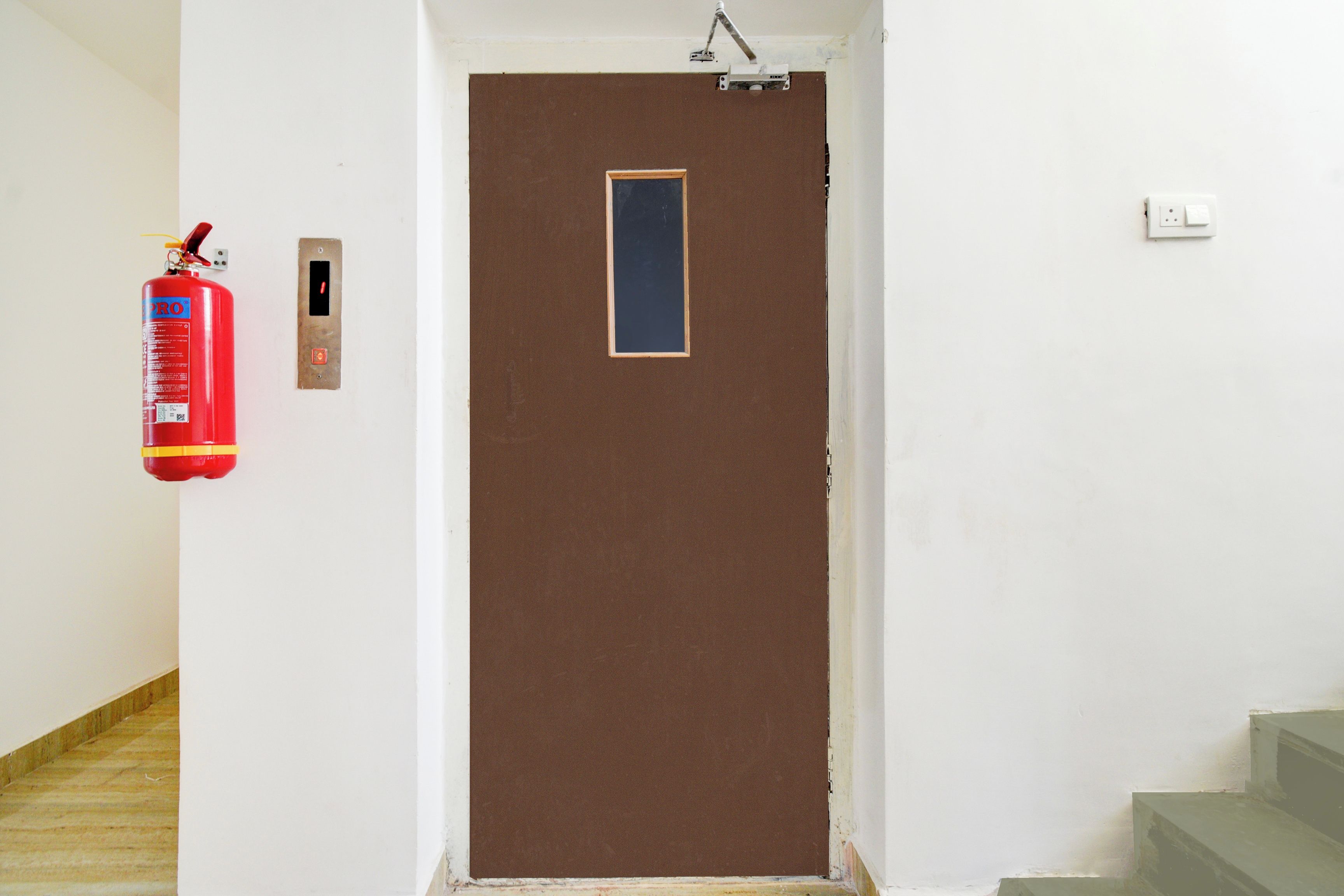 Eine braune Tür mit einem kleinen Fenster, ein an der Wand links angebrachter Feuerlöscher und ein Lichtschalter an der Wand rechts.