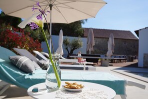 Cenário à beira da piscina com cadeiras de descanso, uma mesa com um prato de biscoitos, um vaso com flores e uma garrafa de água.