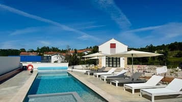 Una piscina con sillones de descanso y sombrillas, un edificio de techo rojo y un cielo azul despejado.