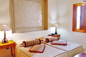 Ein Schlafzimmer mit zwei Betten, jeweils mit Kissen und Lampe ausgestattet. Ein Fenster mit Rollläden und eine Holztür sind ebenfalls vorhanden.